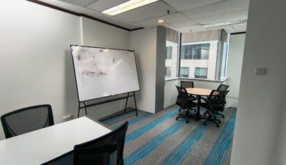 A small office room with a whiteboard, two tables, several black chairs, and large windows letting in natural light.