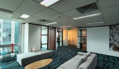 Modern office lounge with grey sofas, a round wooden coffee table, blue carpet, glass doors, and large windows overlooking adjacent buildings.
