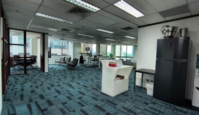 Open office space with blue carpet, workstations, lounge chairs, a fridge, water dispenser, and large windows providing natural light.