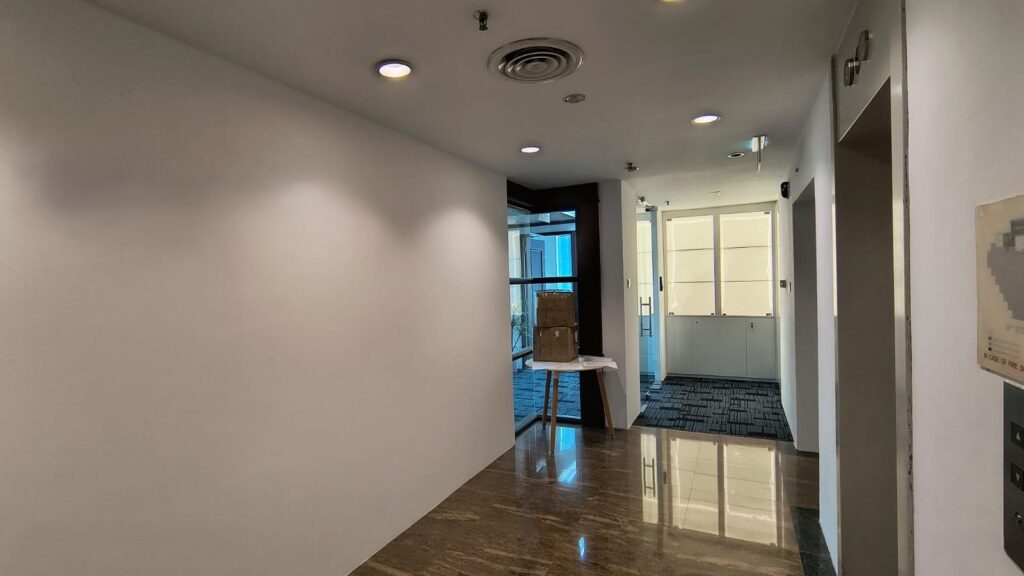 A corridor with white walls, marble flooring, ceiling lights, and doorways leading to rooms with glass panels and carpeted floors. A box sits on a small table near the end of the corridor.