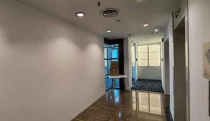 A corridor with white walls, marble flooring, ceiling lights, and doorways leading to rooms with glass panels and carpeted floors. A box sits on a small table near the end of the corridor.