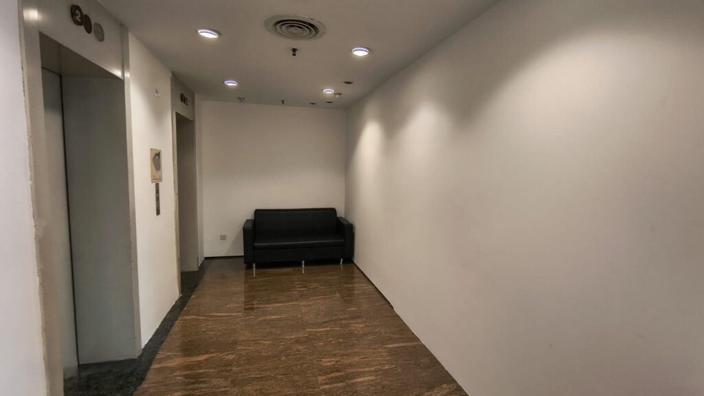 A small lobby area with a black sofa against the back wall, a lift on the left, white walls, and brown tiled flooring.