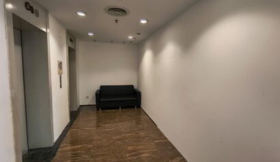 A small lobby area with a black sofa against the back wall, a lift on the left, white walls, and brown tiled flooring.