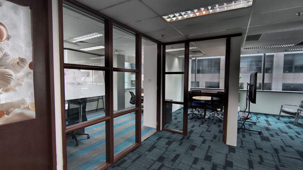 Modern office space with glass-walled meeting rooms, blue and grey carpet, desks, chairs, and large windows letting in natural light.