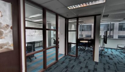 Modern office space with glass-walled meeting rooms, blue and grey carpet, desks, chairs, and large windows letting in natural light.