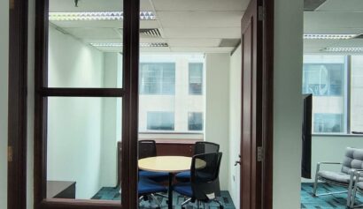 A small office meeting room with a round table, four chairs, glass walls, and an open wooden door, with windows overlooking other buildings.