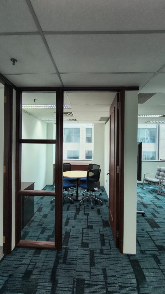 A small office meeting room with a round table, four chairs, glass walls, and an open wooden door, with windows overlooking other buildings.