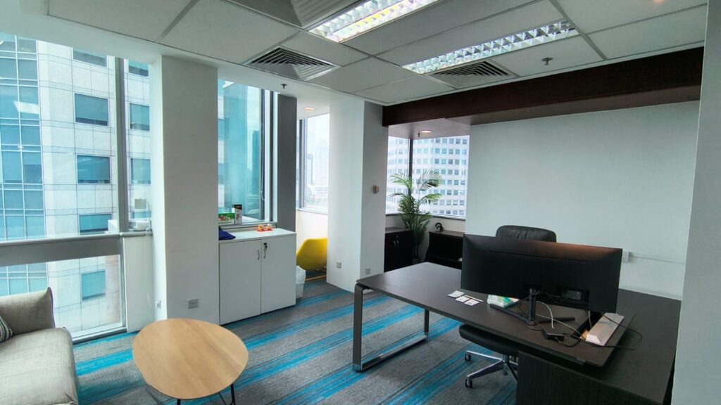 Modern office with a desk, chair, computer, sofa, round table, and shelves by large windows overlooking city buildings.