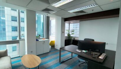 Modern office with a desk, chair, computer, sofa, round table, and shelves by large windows overlooking city buildings.