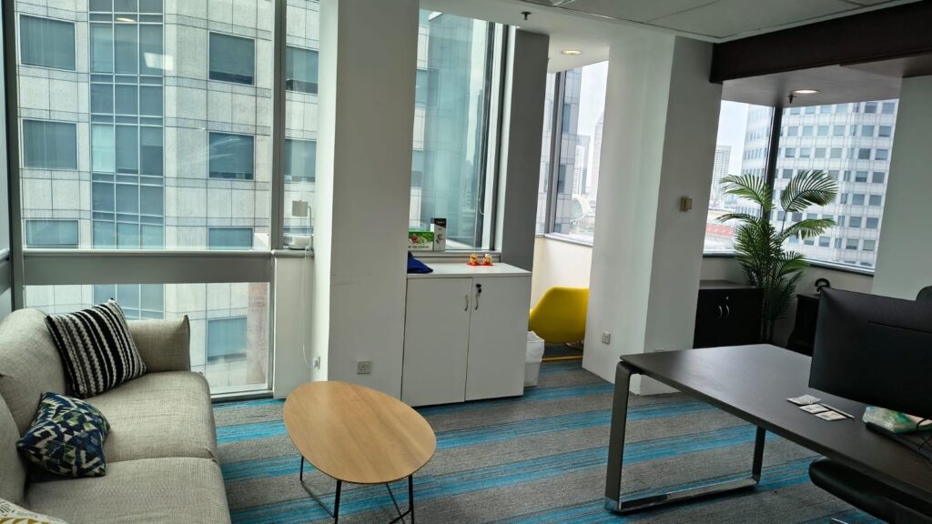 Modern office with large windows, a sofa, round coffee table, desk, cupboard, and a small yellow chair in a corner nook.