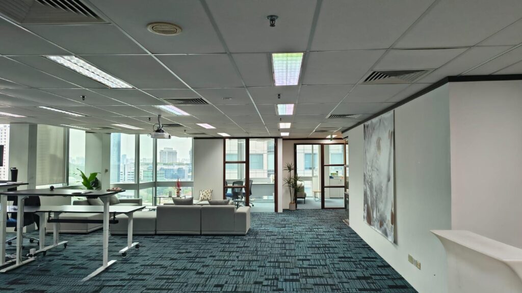 Modern office space with blue carpet, workstations, sofas, large windows, and glass-walled meeting rooms. A large artwork hangs on the right wall.