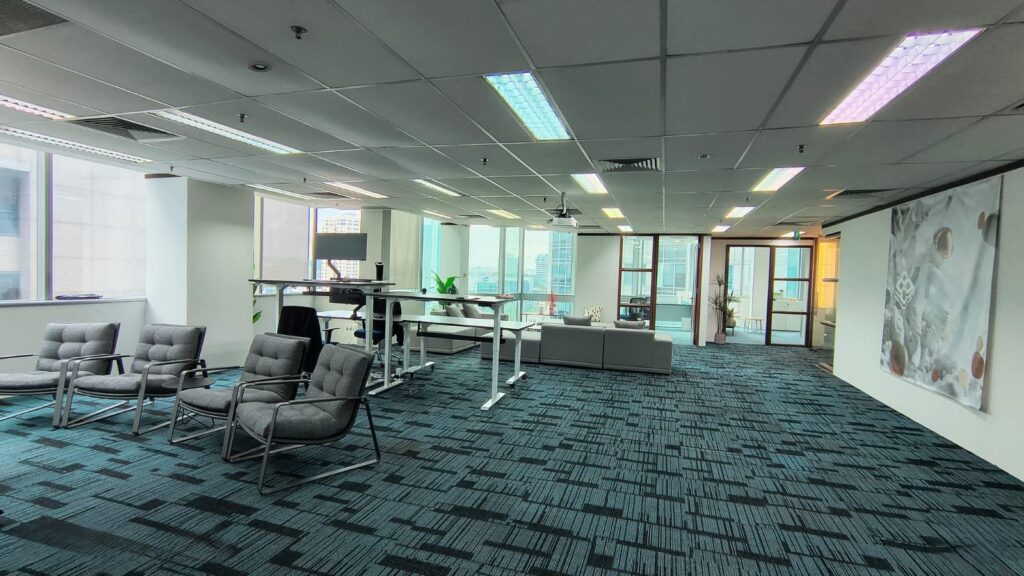 Modern office space with carpeted floors, lounge chairs, work desks, sofas, large windows, and glass doors leading to another room.