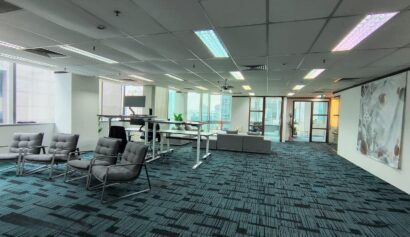 Modern office space with carpeted floors, lounge chairs, work desks, sofas, large windows, and glass doors leading to another room.