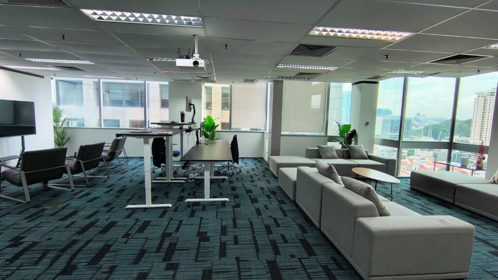Modern office space with large windows, grey sectional sofa, standing desks, lounge chairs, indoor plants, and city views in the background.