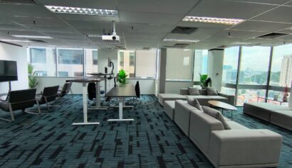 Modern office space with large windows, grey sectional sofa, standing desks, lounge chairs, indoor plants, and city views in the background.