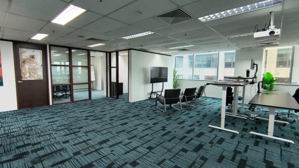 Modern office space with blue carpet, glass-walled meeting room, several chairs, a large television, standing desks, and potted plants by the window.