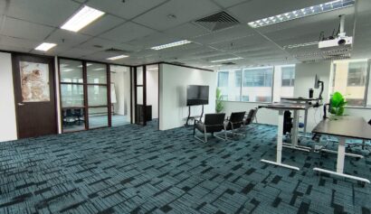Modern office space with blue carpet, glass-walled meeting room, several chairs, a large television, standing desks, and potted plants by the window.