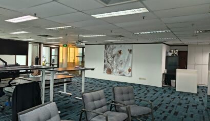 Modern office space with blue carpet, grey chairs, white desks, and a large abstract artwork on the wall. Ceiling lights and glass-walled rooms are visible in the background.