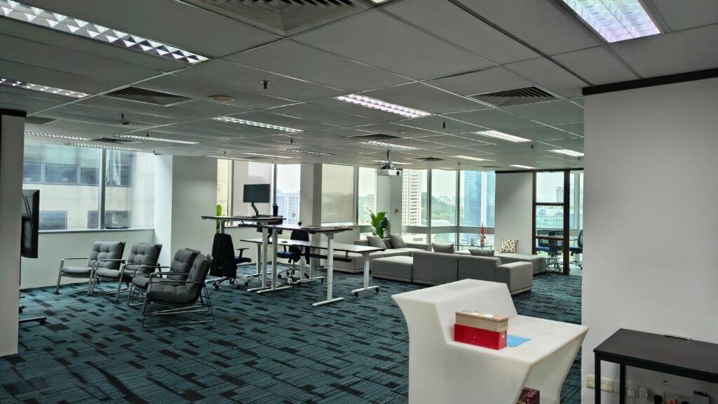 Modern office with large windows, several desks, lounge chairs, sofas, and a high table; blue carpet and ceiling lights visible.