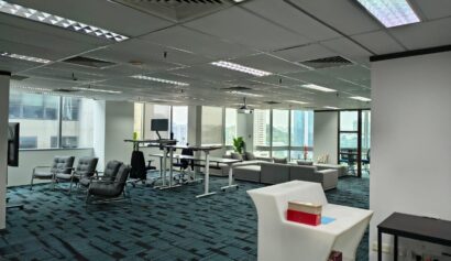 Modern office with large windows, several desks, lounge chairs, sofas, and a high table; blue carpet and ceiling lights visible.