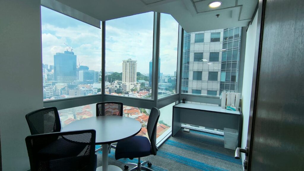 Small office with a round table, four chairs, desk, and large windows overlooking a cityscape with tall buildings and blue sky.