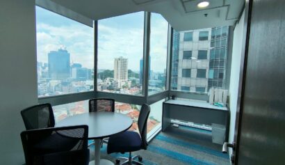 Small office with a round table, four chairs, desk, and large windows overlooking a cityscape with tall buildings and blue sky.