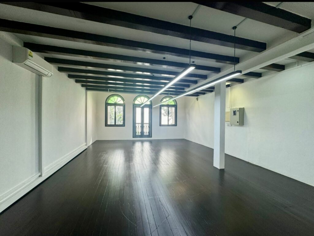 Empty room with dark wooden floors, white walls, exposed ceiling beams, three arched windows, and a door with a small balcony.