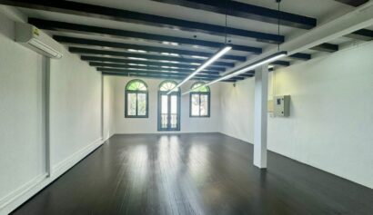 Empty room with dark wooden floors, white walls, exposed ceiling beams, three arched windows, and a door with a small balcony.