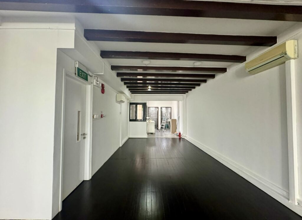 A long, empty room with dark wooden floor and ceiling beams, white walls, air conditioners, and an exit door on the left. Some equipment is visible at the far end.