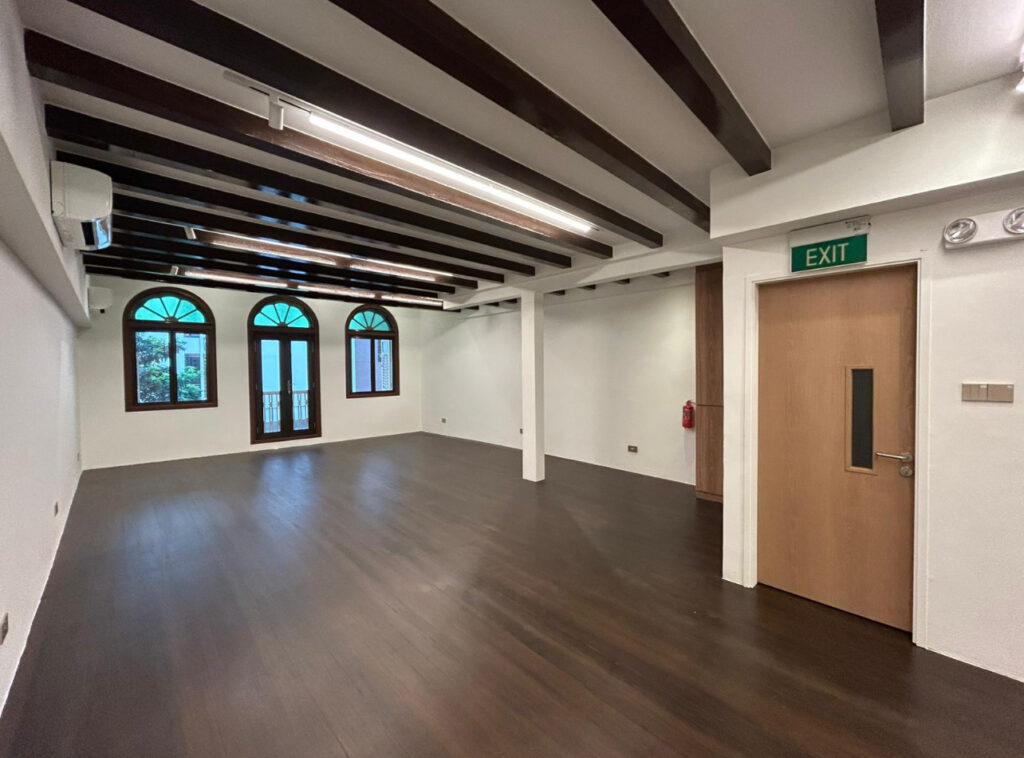 Empty room with dark wooden floors, exposed ceiling beams, white walls, arched windows and doors, an EXIT sign above a wooden door, and a fire extinguisher on the wall.