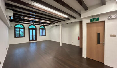 Empty room with dark wooden floors, exposed ceiling beams, white walls, arched windows and doors, an EXIT sign above a wooden door, and a fire extinguisher on the wall.