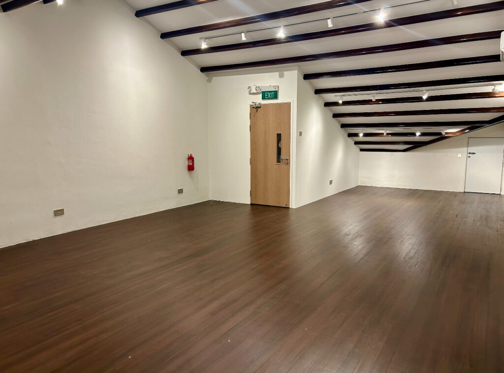 Empty room with wooden floor, white walls, exposed dark ceiling beams, a closed door with an exit sign, and a fire extinguisher mounted on the wall.