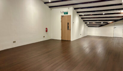 Empty room with wooden floor, white walls, exposed dark ceiling beams, a closed door with an exit sign, and a fire extinguisher mounted on the wall.
