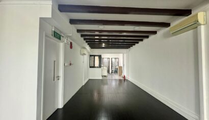 A long, empty room with dark wooden floor and ceiling beams, white walls, air conditioners, and an exit door on the left. Some equipment is visible at the far end.