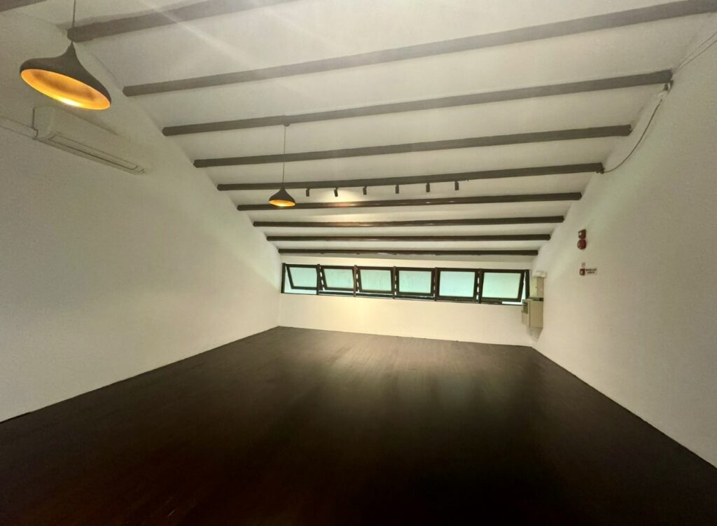 An empty room with dark wooden floors, white walls, exposed ceiling beams, hanging lights, and multiple rectangular windows along one slanted wall.