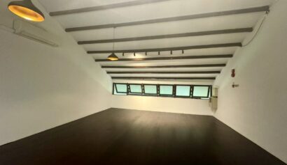 An empty room with dark wooden floors, white walls, exposed ceiling beams, hanging lights, and multiple rectangular windows along one slanted wall.