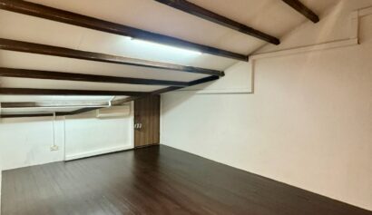 Room with sloping ceiling, exposed wooden beams, white walls, and dark wooden floor; fluorescent light fitting on ceiling.