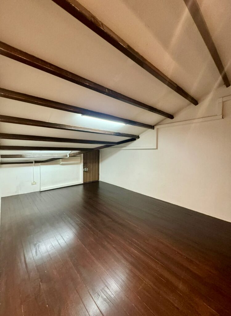 Room with sloping ceiling, exposed wooden beams, white walls, and dark wooden floor; fluorescent light fitting on ceiling.