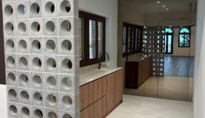 A modern kitchen with a concrete breeze block partition, wooden cupboards, beige floor tiles, and a mirrored wall reflecting the space.
