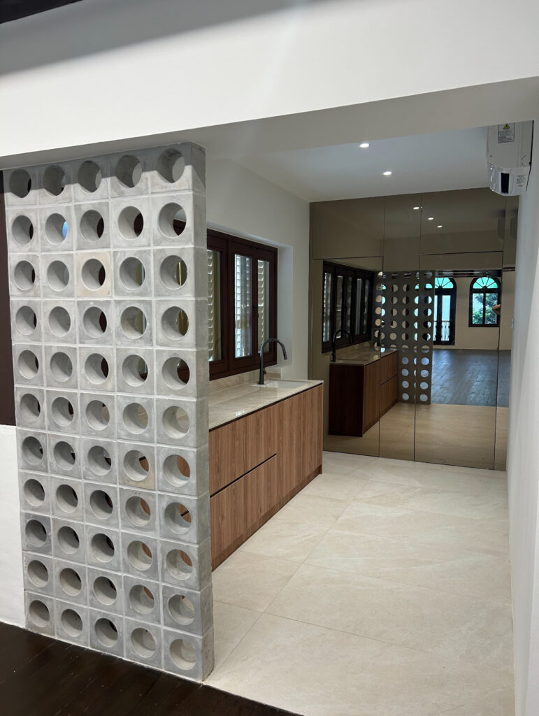 A modern kitchen with a concrete breeze block partition, wooden cupboards, beige floor tiles, and a mirrored wall reflecting the space.