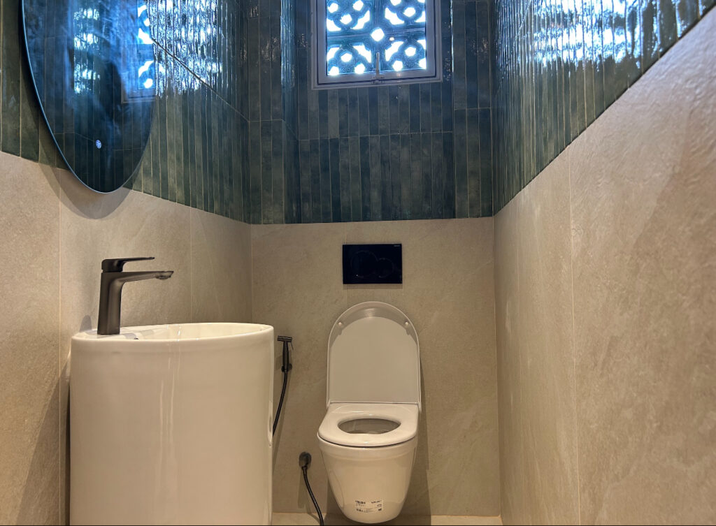 A small bathroom with a white toilet, a modern basin with a single tap, and green tiles on the upper walls beneath a patterned window.