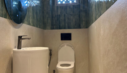 A small bathroom with a white toilet, a modern basin with a single tap, and green tiles on the upper walls beneath a patterned window.
