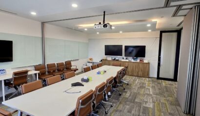 Modern conference room with two long tables, brown chairs, large screens, a projector on the ceiling, and glass walls. The room is well-lit with overhead lighting.