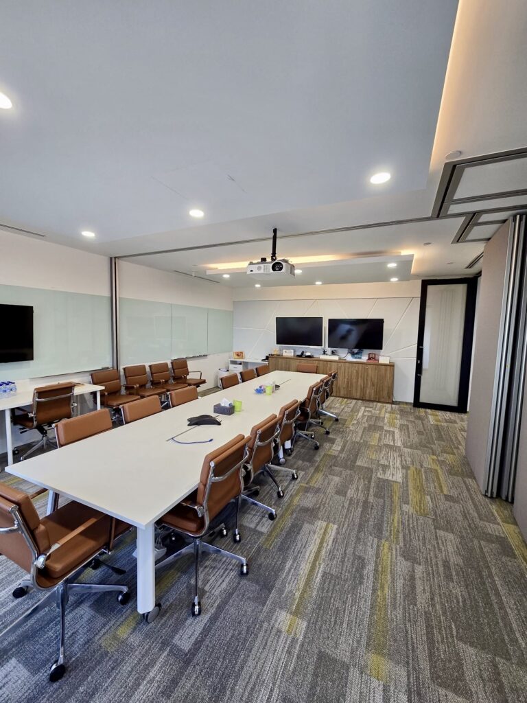 Modern conference room with two long tables, brown chairs, large screens, a projector on the ceiling, and glass walls. The room is well-lit with overhead lighting.