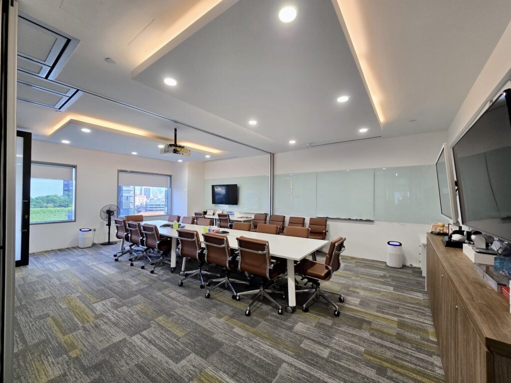 Modern conference room with a long white table, brown office chairs, large windows, wall-mounted screens, and a whiteboard. The room has bright lighting and a contemporary design.
