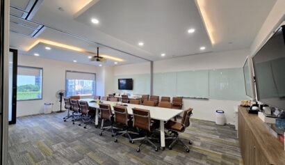 Modern conference room with a long white table, brown office chairs, large windows, wall-mounted screens, and a whiteboard. The room has bright lighting and a contemporary design.