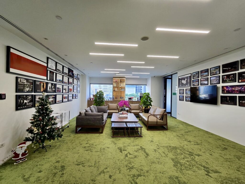 Modern office lounge with green carpet, sofas, coffee tables, wall art, and a small decorated Christmas tree; large windows in the background.