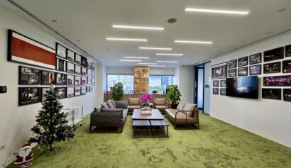 Modern office lounge with green carpet, sofas, coffee tables, wall art, and a small decorated Christmas tree; large windows in the background.