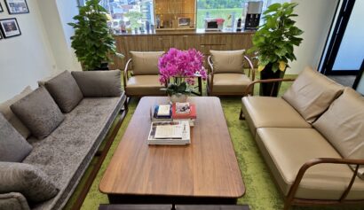 Modern office lounge with sofas, a wooden coffee table with books and flowers, two stools, potted plants, and large windows showing a cityscape in the background.