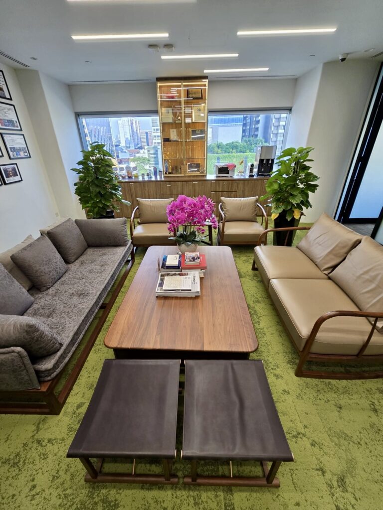 Modern office lounge with sofas, a wooden coffee table with books and flowers, two stools, potted plants, and large windows showing a cityscape in the background.